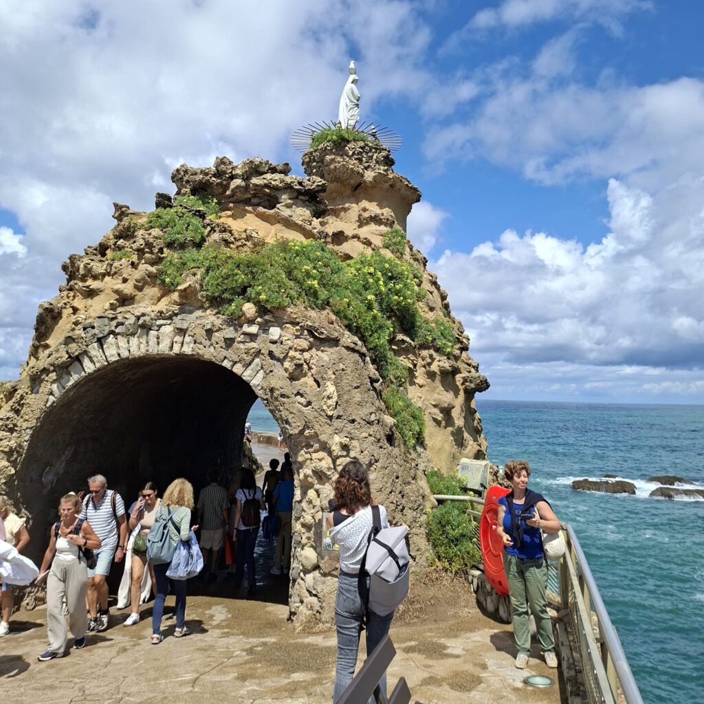 Egy Szűz Mária-szobor egy kis sziklán, a sziklában alagút, amelyen emberek járnak át, a háttérben a tenger