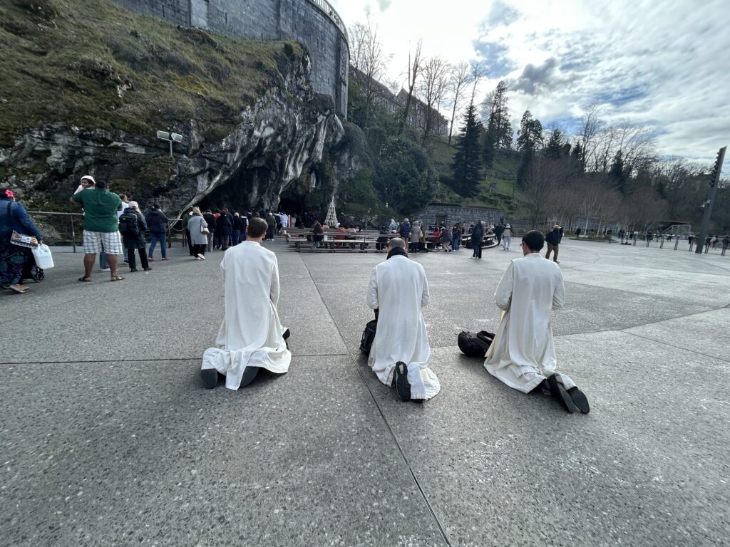 Három férfi fehér albában térdel Lourdes-ban a barlang előtt